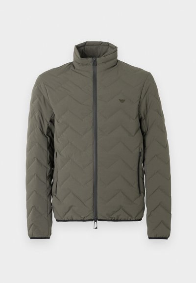 Olivovozelená prešívaná bunda s vysokým golierom, predným zipsom a vzorom šípok. Má čierne detaily pozdĺž zipsu a manžiet.