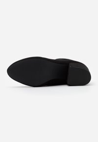 Zapato de tacón alto de ante negro con una suela texturizada, presentando una forma curva y suave y un tacón en bloque. La suela es plana y está hecha de goma.