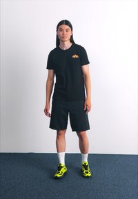 Crna majica s narančastim logom, tamnoplave traper kratke hlače i neonske žute tenisice. Model stoji na sivoj prostirci ispred jednostavne bijele pozadine.