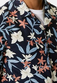 Kortärmad skjorta i marinblått tyg med blommönster i vitt, rosa och blått, kompletterat med gröna blad och en avslappnad krage.