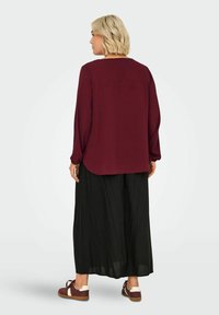 Burgundy long-sleeve blouse with a rounded hem, paired with black wide-leg trousers and brown and white sneakers. Smooth texture, relaxed fit.