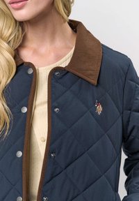 Vrouw draagt een marineblauwe doorgestikte jas met een bruine corduroy kraag over een crèmekleurig topje met knopen, met een geborduurd paard en ruiterlogo op de borst.