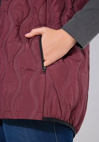 Burgunderfarbene, gesteppte Weste mit gewellter Textur, ausgestattet mit einer Reißverschlusstasche an der Seite und einem elastischen Saum. Getragen über einem grauen langärmligen Oberteil und blauen Jeans.