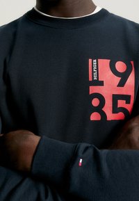 Persona que lleva una sudadera azul oscuro con el logotipo rojo y blanco "Hilfiger 1985" en el pecho y un pequeño emblema de bandera en el puño de la manga.