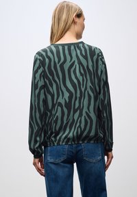 Maglietta a maniche lunghe di colore verde scuro con strisce zebra nere, caratterizzata da una vestibilità rilassata e un colletto rotondo, abbinata a jeans di denim blu.
