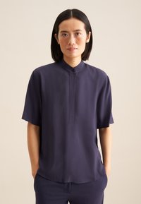 Camicia corta in navy con colletto alto, patta abbottonata e tessuto liscio. Presenta una vestibilità comoda e una leggera lucentezza.