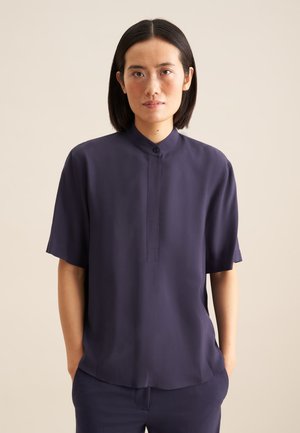Camicia corta in navy con colletto alto, patta abbottonata e tessuto liscio. Presenta una vestibilità comoda e una leggera lucentezza.