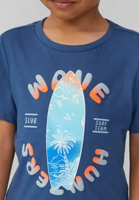 Blaues Baumwoll-T-Shirt mit einem Surfbrett-Graphic, das eine Palme und eine Ozeanszene zeigt, umgeben von dem Text "WAVE SURF TEAM" in Weiß und Orange.