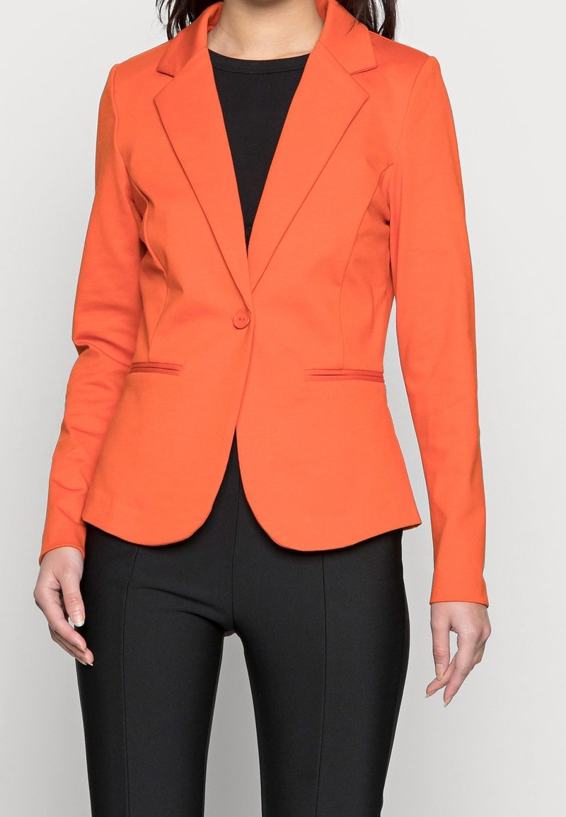 Femme portant un blazer orange ajusté à un seul bouton sur un haut et un pantalon noirs, debout devant un fond clair uni.