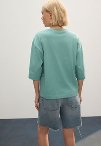 Tricou larg, de culoare verde deschis, cu mâneci 3/4. Asortat cu pantaloni scurți din denim deschis, cu margini destrămate și croială lejeră.