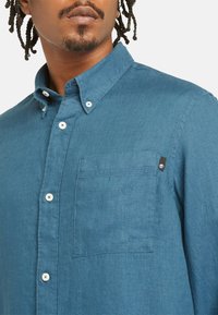 Camisa verde-água abotoada, feita de tecido suave, com um bolso no peito e botões brancos. O colarinho tem um estilo clássico com costura visível.