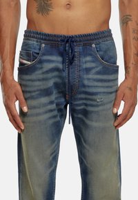 Joggers en denim avec un dégradé de lavage, taille élastique, cordon de serrage, poches latérales et détails d'usure subtils sur le tissu.