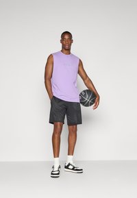 Chemise sans manches violet clair avec logo, shorts en mesh noirs, et baskets noir et blanc. Tenant un ballon de basket. Fond simple.