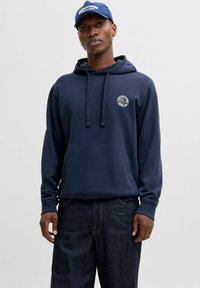 Navy blue hoodie made of soft fabric, featuring a front pocket, adjustable drawstring hood, and a small logo on the chest. Worn with dark jeans.