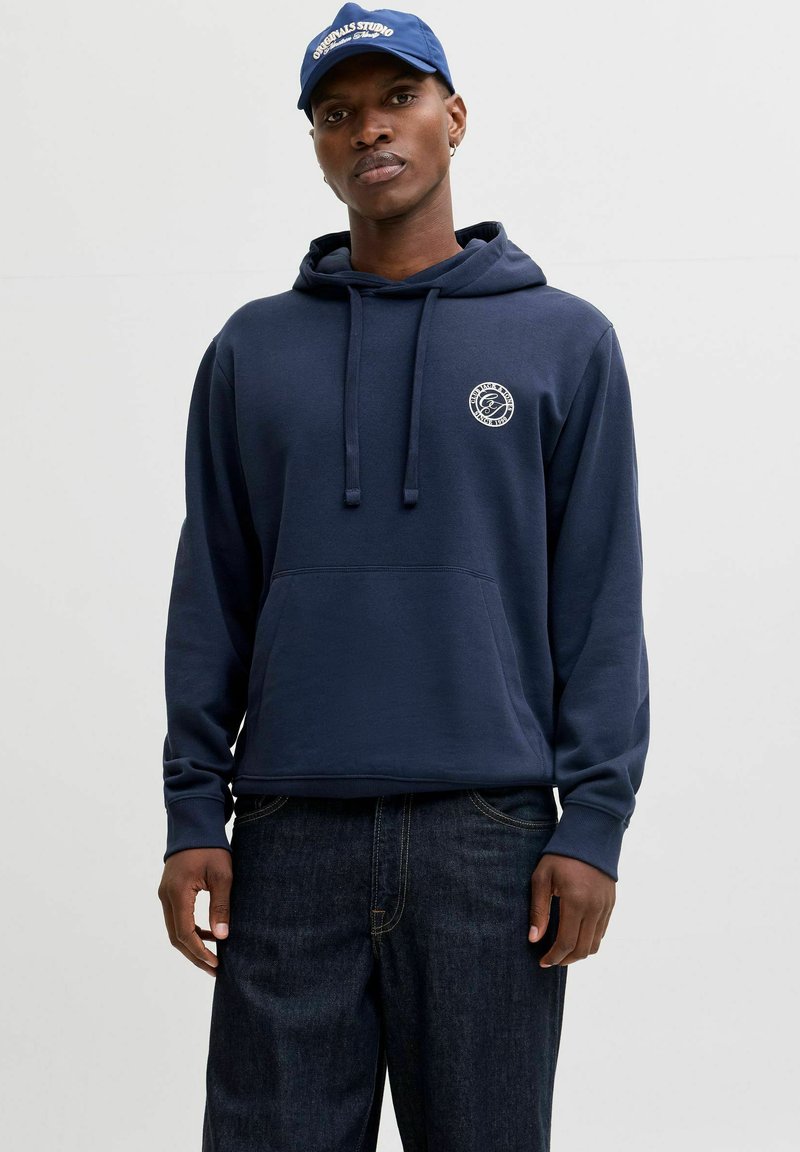 Navy blue hoodie made of soft fabric, featuring a front pocket, adjustable drawstring hood, and a small logo on the chest. Worn with dark jeans.