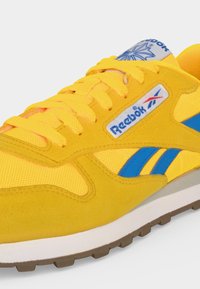 Κίτρινο αθλητικό παπούτσι Reebok με μπλε ρίγες λογότυπου, λευκή ενδιάμεση σόλα, καφέ εξωτερική σόλα και κίτρινα κορδόνια σε λευκό φόντο.