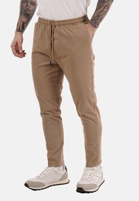 Uomo che indossa pantaloni beige slim-fit con vita regolabile e sneakers bianche su sfondo bianco.