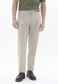 Pantaloni beige chiaro in tessuto texture, con pieghe frontali singole e chiusura a bottone, abbinati a scarpe stringate marroni.