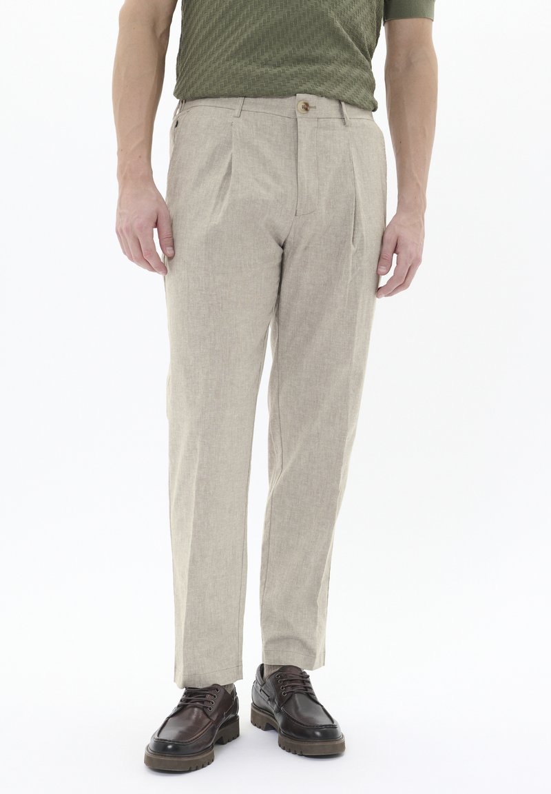 Pantaloni beige chiaro in tessuto texture, con pieghe frontali singole e chiusura a bottone, abbinati a scarpe stringate marroni.