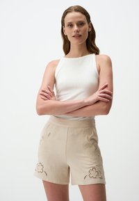 Beige Shorts mit floralem Stickerei und Ausschnitten, kombiniert mit einem figurbetonten weißen Tanktop. Glattes Material mit lockerer Passform.