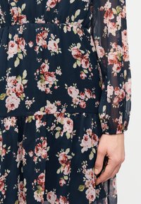 Το χέρι μιας γυναίκας ακουμπά στη μέση σε φόρεμα από διαφανές ύφασμα σκούρου μπλε χρώματος με ροζ και πράσινο λουλουδένιο μοτίβο και λεπτομέρεια με σούρες στη μέση.