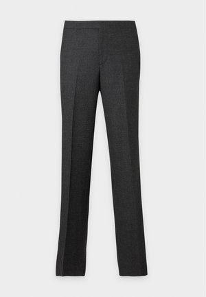 Homme portant un pull polo gris clair à manches longues, un pantalon gris foncé et des chaussures noires, debout avec une main dans la poche sur un fond blanc.