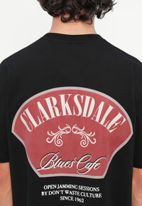 Camiseta de algodón negra con un gran gráfico en rojo y blanco en la parte trasera que incluye el texto "CLARKSDALE Blues Cafe" y acentos decorativos.