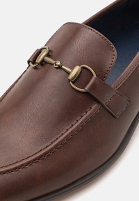 Bruna läderloafers med rund tå, dekorativt bronsbeslag och synlig söm längs kanten. Slät yta med mattslät finish.