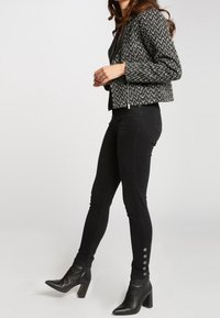 Svart kort jacka med ett fiskbensmönster, i kombination med åtsittande svarta byxor med knappramar vid vristerna och klackade svarta stövlar.