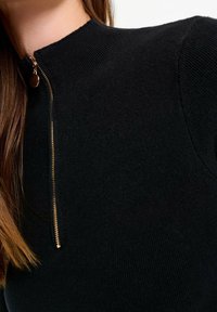 Svart stickad topp med hög krage och frontdragkedja med ett guldfäste. Texturen är ribbad och sitter åtsittande på kroppen.