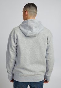 Sudadera gris de tejido suave, con capucha ajustable, puños y dobladillo acanalados. Mostrada desde la parte de atrás con detalles mínimos.