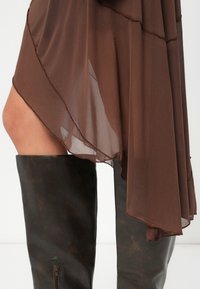 Καφέ διαφανής φούστα με στρωτό στρίφωμα που κρέμεται πάνω από μποτάκια από σκούρο καφέ δέρμα μέχρι το γόνατο, με φερμουάρ στο πλάι. Λείος υφασμάτινος ιστός και γυαλιστερό φινίρισμα.