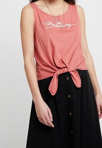 Koralfarvet tanktop med knude foran, med hvid "Billabong"-logo. Båret med en sort nederdel med knapper ned foran.