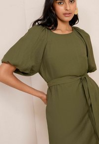 Rochie verde măsliniu cu mâneci scurte bufante, țesătură texturată, decolteu rotund și curea legată în talie. Detalii de design simple și elegante.