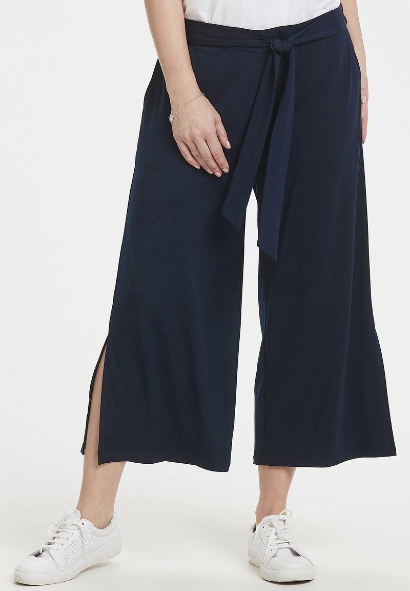 Pantaloni a gamba larga blu navy realizzati in tessuto morbido, con una cintura annodata e spacchi laterali, abbinati a sneakers bianche.