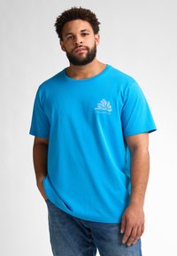 Tepaná modrá bavlněná tričko s krátkým rukávem a kulatým výstřihem, se tištěným logem na levé hrudi. Nosené s džíny, které mají volný střih.