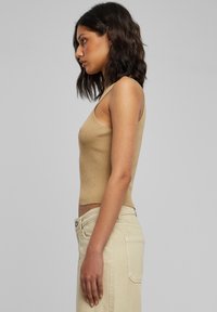 Kvinde med skulderlangt mørkt hår iført en ærmeløs beige ribbet top og lyse beige bukser, stående i profil mod en ensfarvet baggrund.