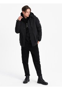 Uomo vestito con giacca invernale nera con cappuccio, pantaloni cargo neri, felpa nera con cappuccio e stivali neri stringati, in piedi su sfondo bianco.