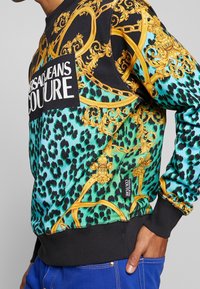 Sudadera en negro, verde y oro con un estampado de leopardo y un patrón barroco; cuenta con un parche de logo y puños y dobladillo acanalados.