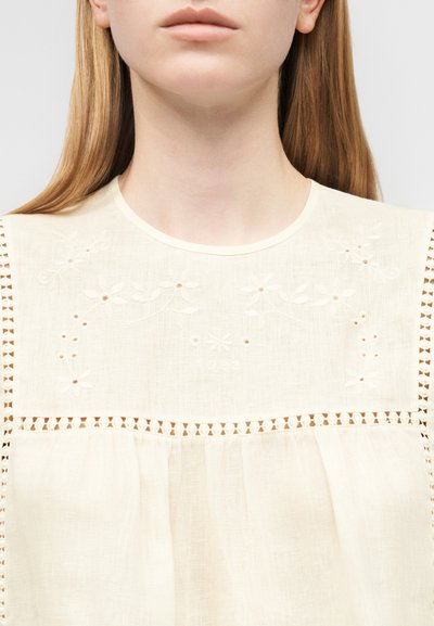 Κοντινό πλάνο γυναίκας που φοράει μπεζ αμάνικη μπλούζα με λουλουδάτη κέντηση και δαντέλα γύρω από το στήθος και τους ώμους.