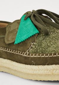 Chaussure en daim vert avec une tige texturée, des détails cousus et une semelle en jute tissé. Équipée d'une étiquette bleu-vert avec "CLARKS ORIGINALS" embossé.