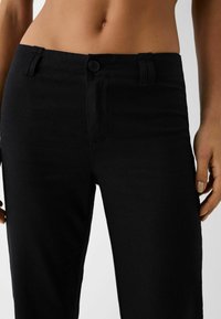 Black linen trousers feature a button closure, side pockets, and a straight-leg design. The fabric has a smooth texture and subtle sheen.