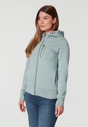 Chaqueta con capucha y cremallera en color verde claro, fabricada en tela suave con un corte ajustado. Presenta una cremallera frontal, bolsillos y un logo en el pecho.