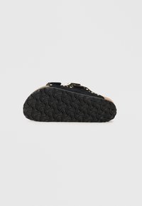 Chaussure en daim noir avec une semelle en caoutchouc texturée, ornée de clous dorés et d'un accent en liège. Sangle ajustable pour un maintien sûr.