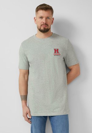 Man met baard, gekleed in een lichtgrijs T-shirt van Harvard University en blauwe spijkerbroek, staand tegen een effen lichte muur.