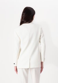 Hvit skreddersydd blazer med teksturert stoff, med en enkelt slisse bak og svarte knapper på ermene. Minimalistisk design, strukturert form.