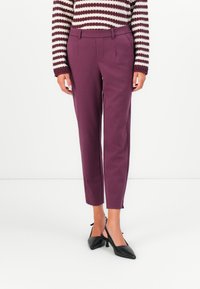 Woman wearing purple tailored ankle pants, black slingback heels, and a maroon-and-white striped knit sweater standing on a white floor.