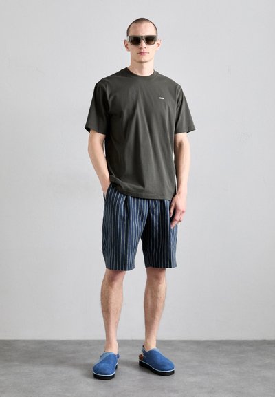 Mørkegrøn kortærmet t-shirt, bl�å stribede shorts og blå slip-on sko. Modellen bærer solbriller og står foran en grå baggrund.