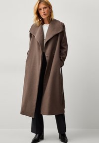 Cappotto lungo in lana marrone con colletto oversize, doppi bottoni, tasche laterali e design dritto, abbinato a pantaloni neri e stivali con tacco.