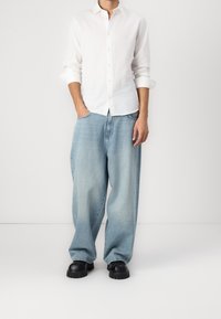 Camicia bianca in lino con bottoni, jeans in denim blu chiaro di taglio morbido, scarpe chunky nere, mani in tasca, sfondo minimalista.
